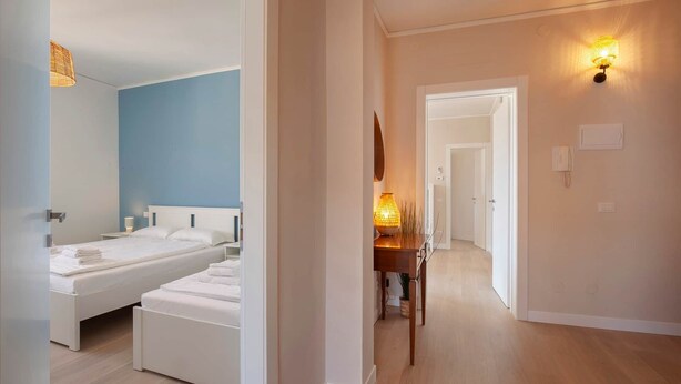 Una habitación de hotel con cama, mesita de noche, lámpara y un pasillo que conduce a otra habitación.