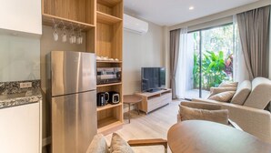 Ein modernes Wohnzimmer mit großem Fernseher, einer Couch, einem Esstisch und einer Küchenzeile inklusive Kühlschrank und Kaffeemaschine.