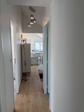 A hallway with a ceiling light fixture, a white door, and a view into a kitchen area with a window.