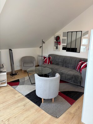 A modern living room with a glass coffee table, grey sofa, and red and white pillows. There is a floor lamp and a tall standing heater.