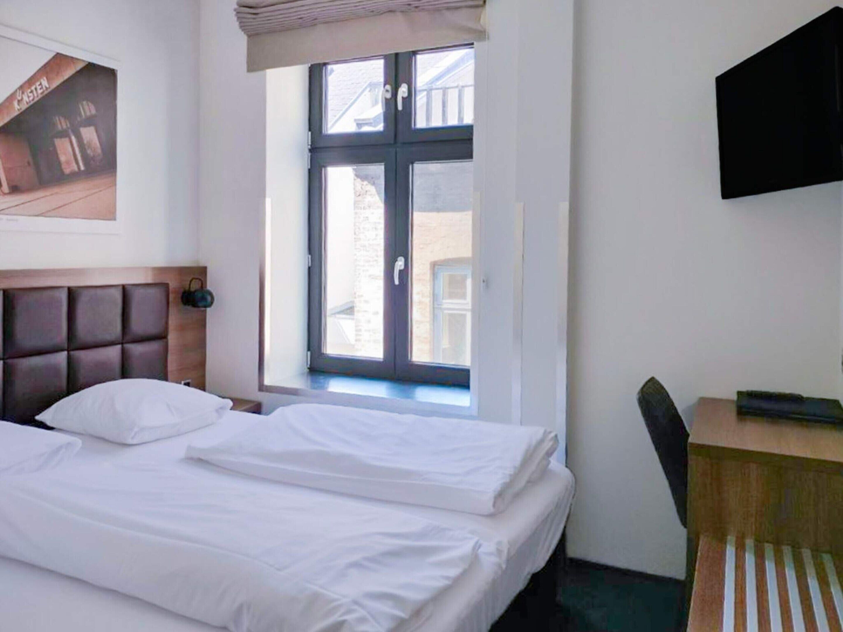 Et hotelværelse med en seng, et skrivebord, et fjernsyn og et vindue med udsigt til bygninger.