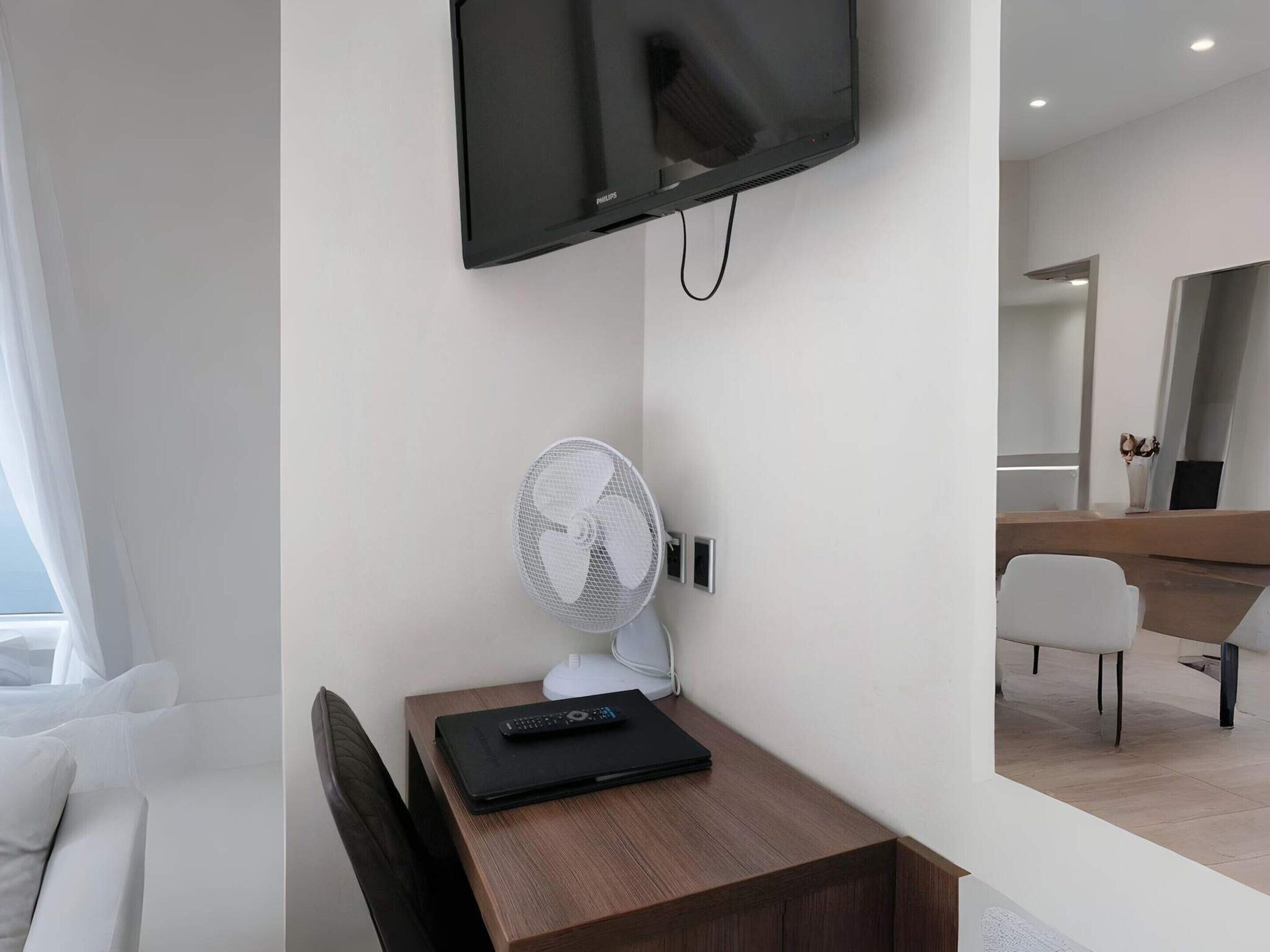 Et hotelværelse med et fladskærms-tv monteret på væggen, en ventilator, et træbord med en fjernbetjening og en hvid stol.