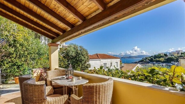 Ein überdachter Außenbereich mit Korbmöbeln und einem Tisch, der auf eine Küstenlandschaft mit Gebäuden und blauem Himmel blickt.
