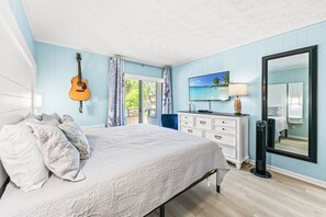 Ein Schlafzimmer mit einem Bett, einer Gitarre, einem Fernseher, einer Kommode, einem Spiegel und einem Fenster.