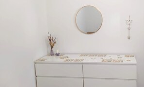 Une commode blanche avec un miroir au-dessus, un petit vase contenant des fleurs séchées et un élément décoratif fixé au mur.