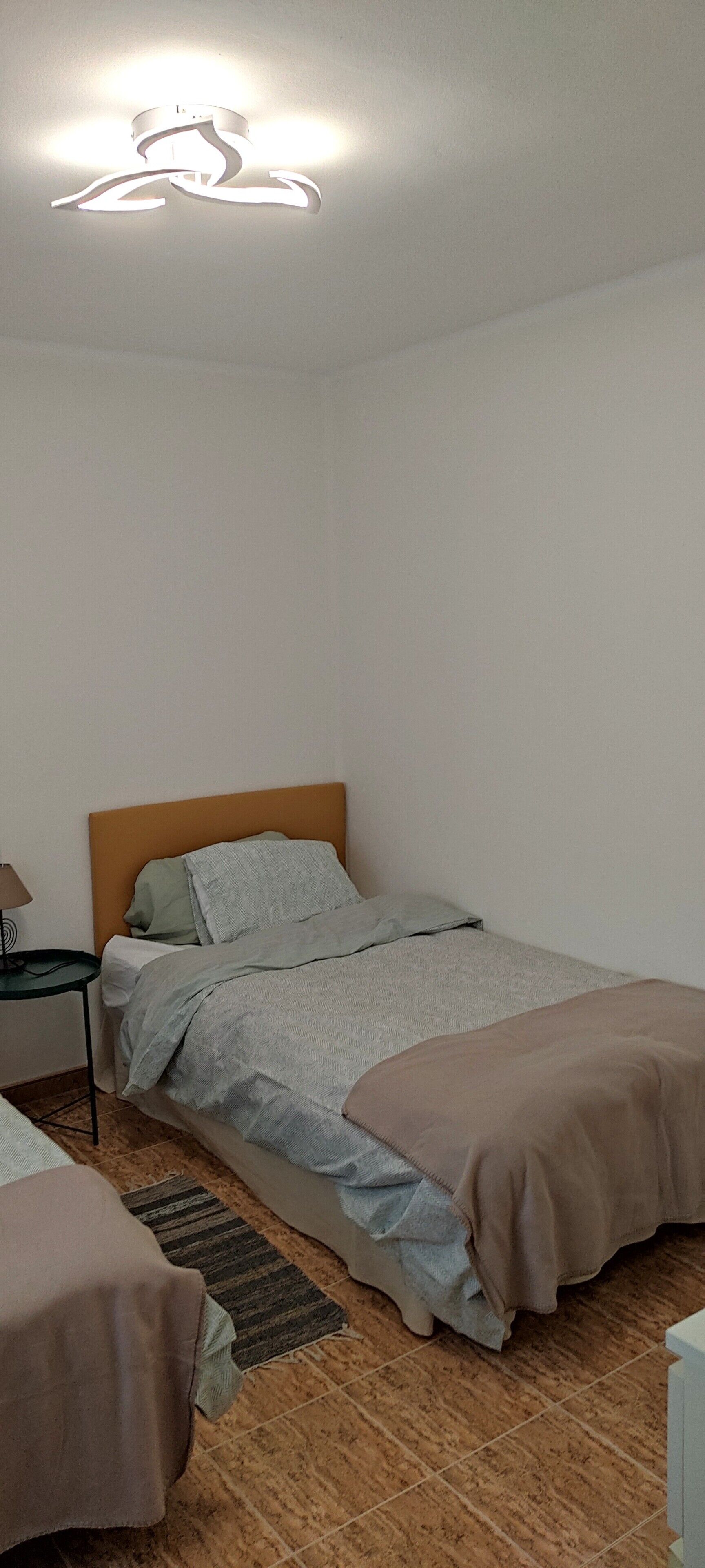 A single bed with a wooden headboard, a bedside table with a lamp, and a patterned rug on a tiled floor.