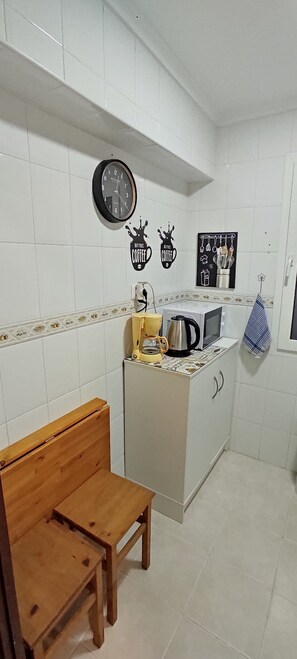 Une petite cuisine avec des carreaux blancs, un banc en bois, un four à micro-ondes, une machine à café et des éléments décoratifs sur les murs, notamment une horloge et des objets sur le thème du café.