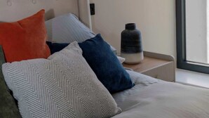 Ett modernt sovrum med en säng, kuddar, ett sängbord, en lampa och ett fönster.