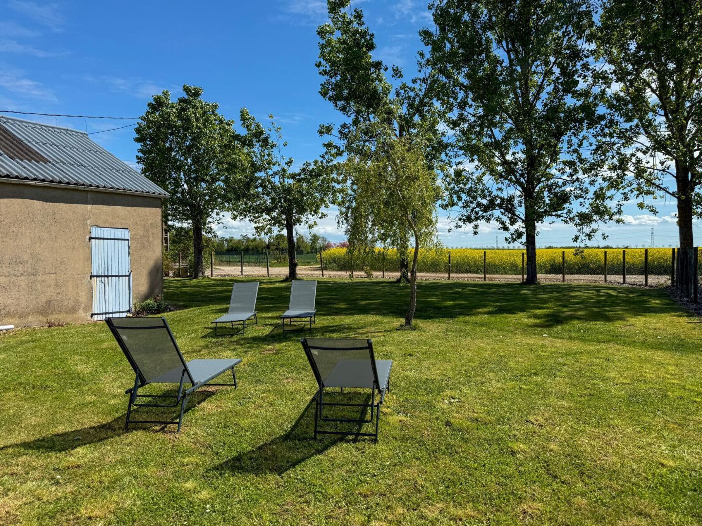 Un espace extérieur verdoyant avec trois chaises longues, un bâtiment doté d’un toit en métal et une clôture en arrière-plan.