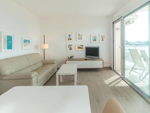 Ein modernes Wohnzimmer mit einer Couch, einem Fernseher auf einem Schrank, einem Tisch und einem Balkon mit Sitzgelegenheiten.