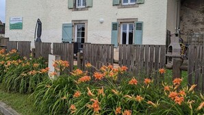 Un bâtiment à l’extérieur blanc, avec des volets verts et une clôture en bois. Des fleurs orange se trouvent au premier plan.
