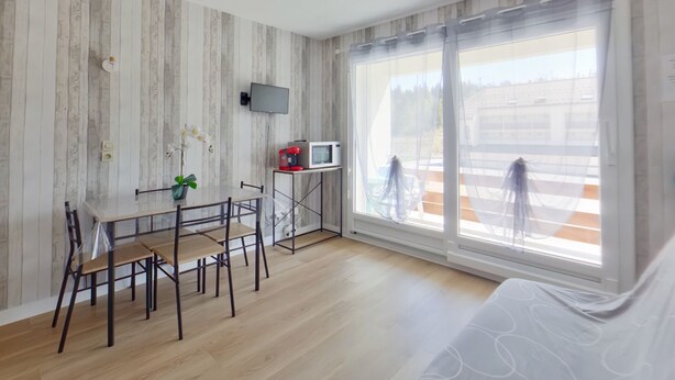 Quarto moderno com piso de madeira, uma mesa de jantar com cadeiras, TV fixada na parede e janelas amplas com vista exterior.