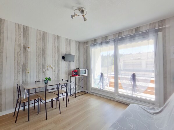 A modern room with a wooden floor, a dining table with chairs, a TV mounted on the wall, and large windows with a view of the outside. - Charming apartment with WiFi - 4 guests (Prémanon)