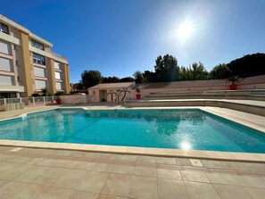 Ein Schwimmbecken mit gefliestem Deck, ein Gebäude mit zahlreichen Fenstern und ein klarer Himmel.