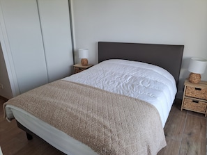 A neatly made bed with a white duvet and a beige throw blanket, a bedside lamp, and a small wooden nightstand with a woven lamp base. - Apartment sea view - 4 guests (Fort-Mahon-Plage)