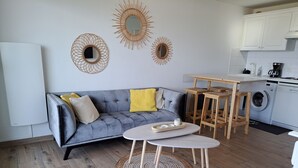 A modern living space with a grey sofa, wooden coffee table, and a kitchen area with a washing machine. - Apartment sea view - 4 guests (Fort-Mahon-Plage)