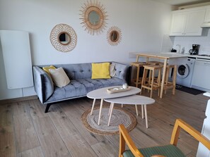 A modern living space with a grey sofa, wooden coffee table, and a kitchen area with a washing machine. - Apartment sea view - 4 guests (Fort-Mahon-Plage)