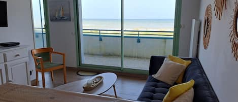 Ein Zimmer mit einer Sitzecke, einem Sessel, einem Tisch, einem Fernseher und Meerblick durch eine Schiebetür.