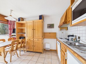 Eine kleine Küche mit Holzausbau, einem Esstisch mit Stühlen, einem eingebauten Backofen und einem Fenster mit Vorhängen.