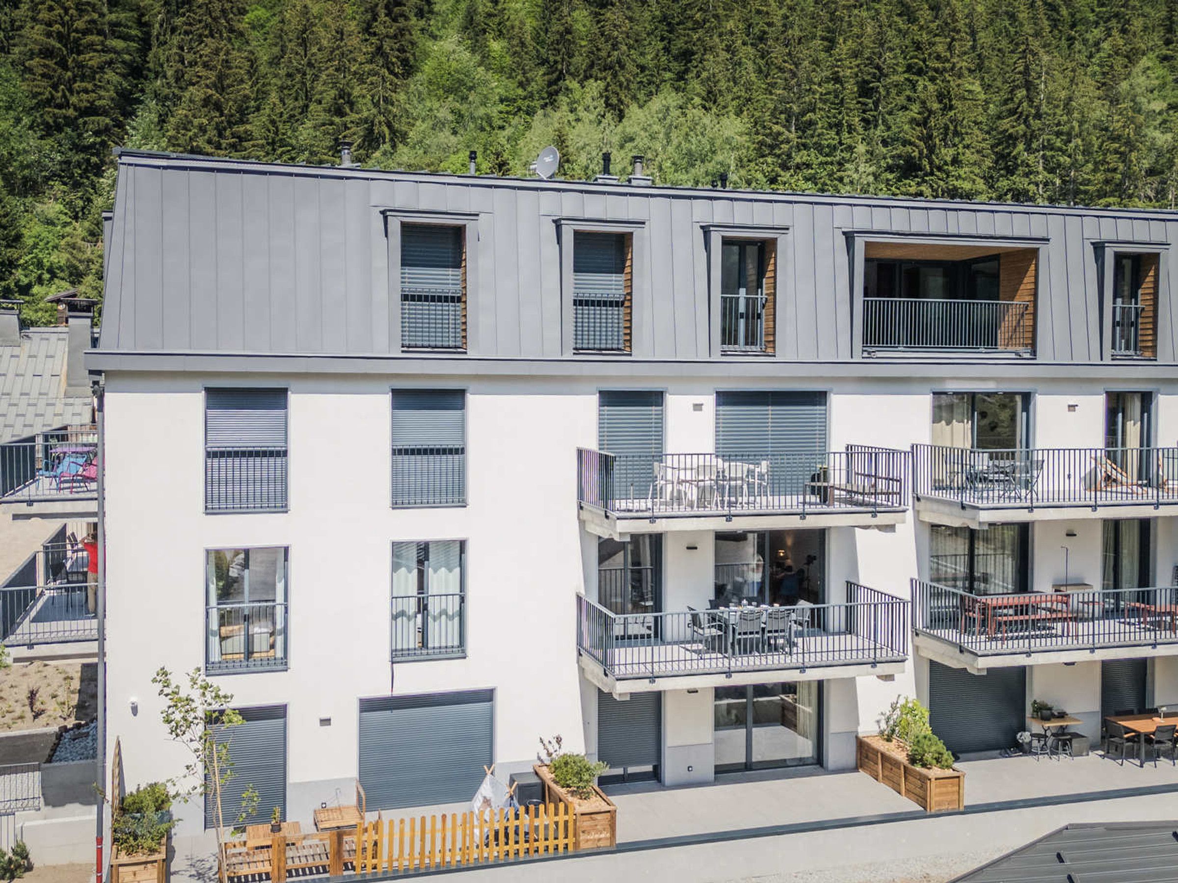A multi-story building with balconies, outdoor seating, and a view of a forested area.
