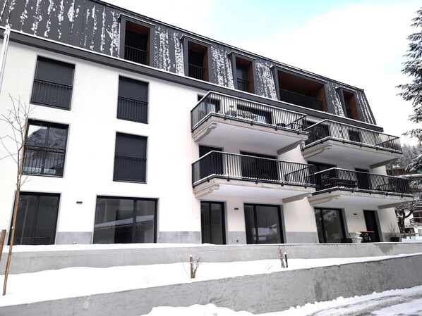 A multi-story building with balconies, large windows, and a snow-covered ground.
