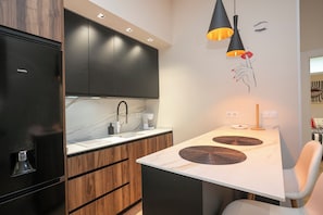 A modern kitchen with a black refrigerator, wooden cabinets, a white countertop, and a dining area with a white chair. - Apartment la serna (Cabezón de la Sal)