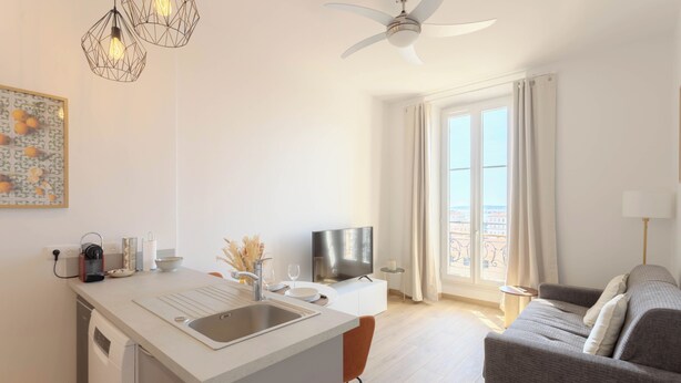 A modern kitchen with a built-in island, a dining area with a sofa, a TV, and a balcony with curtains. - Maison Mon Idée | Top floor, Bright, sea view and Palais des Festivals (Cannes)