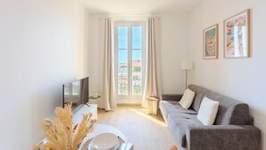 A modern living room with a grey sofa, a flat-screen TV, a coffee table, and a large window with curtains. - Maison Mon Idée | Top floor, Bright, sea view and Palais des Festivals (Cannes)