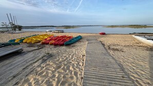 En sandstrand med en träpromenad som leder ner till stilla vatten. Kajaker och kanoter står uppradade på stranden.