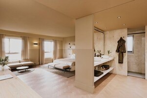 A modern bedroom with a large bed, a bedside table, a desk, and a bathroom with a sink and mirror.