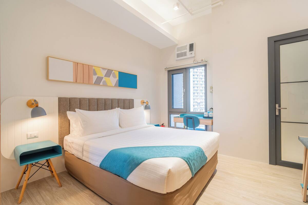 Une chambre d’hôtel avec un lit, une table de chevet, une lampe et une vue sur le paysage urbain par la fenêtre.