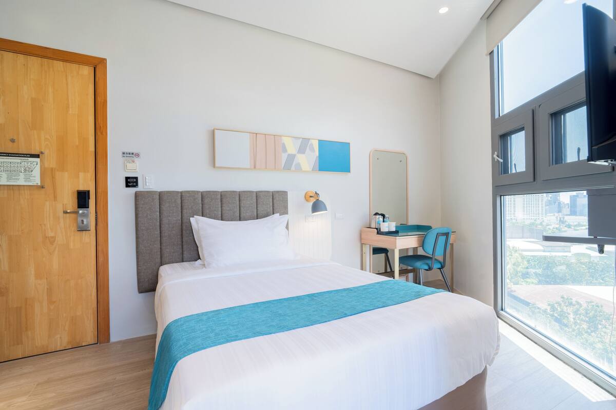 Une chambre d’hôtel avec un lit, un bureau, une chaise, une grande fenêtre et une porte.