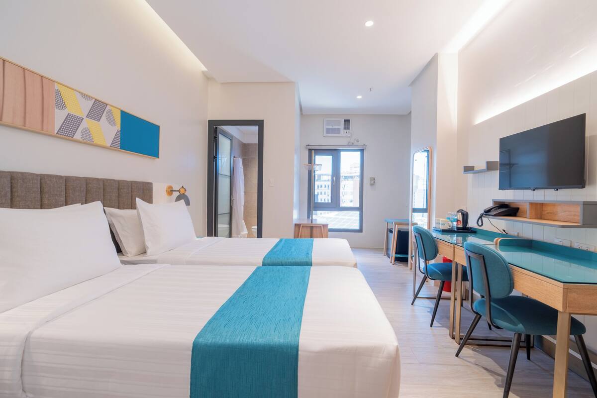 Une chambre d’hôtel avec deux lits, un bureau avec des chaises, une télévision et une fenêtre.