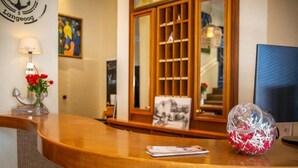 A reception desk with a lamp, a vase with red flowers, a framed picture, and a glass jar with white objects. - HOTEL DÜNENLÄUFER Langeoog (Langeoog)