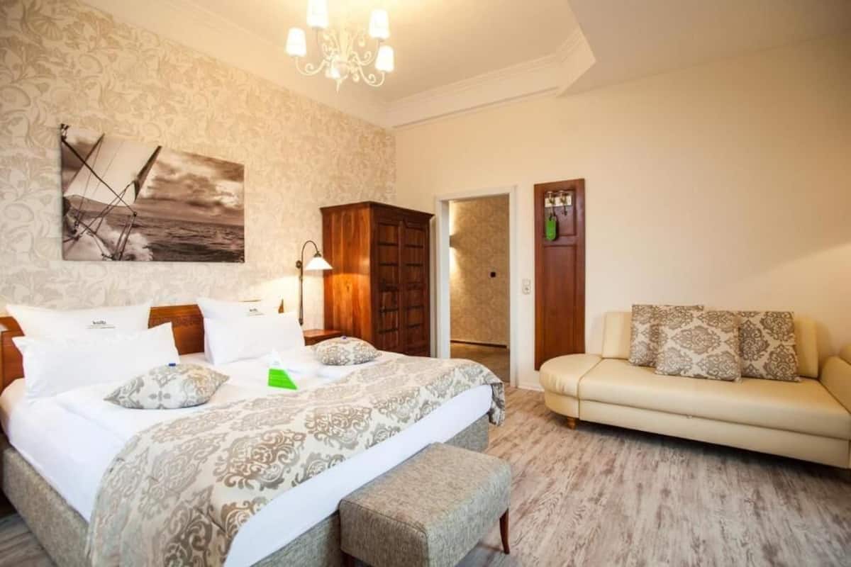 A bedroom with a bed, a bedside table, a lamp, a wooden wardrobe, a door, and a sofa.