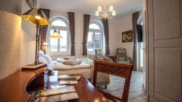 A hotel room with a bed, a desk with papers, a chair, a chandelier, and arched windows. - HOTEL DÜNENLÄUFER Langeoog (Langeoog)