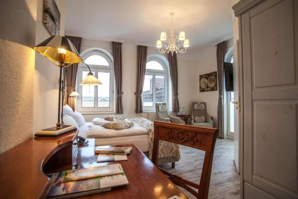 A hotel room with a bed, a desk with papers, a chair, a chandelier, and arched windows.