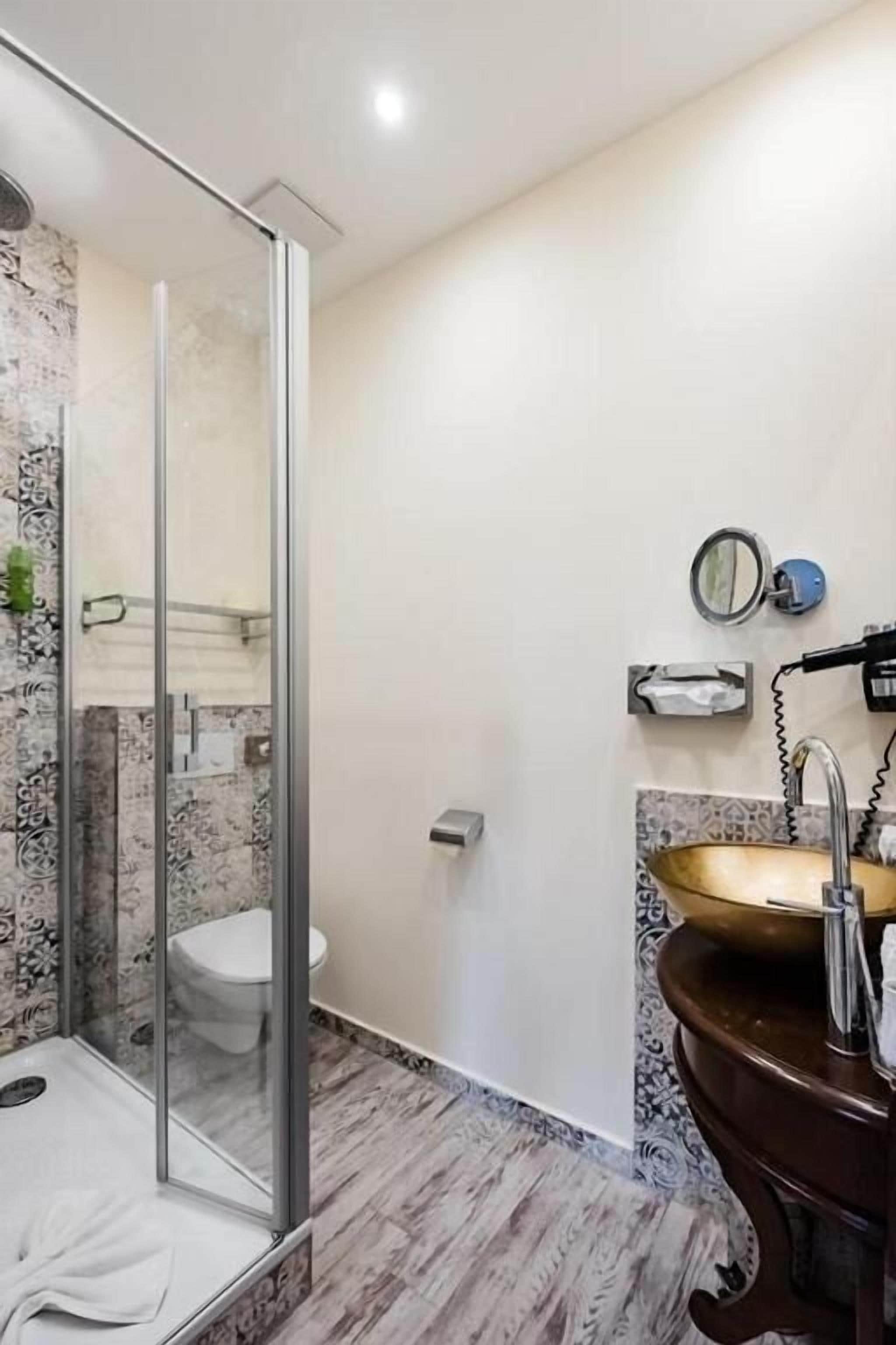 A bathroom with a glass shower enclosure, a marble-tiled wall, a wooden vanity with a gold sink, and a toilet.