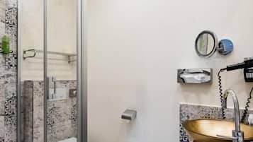 A bathroom with a glass shower enclosure, a marble-tiled wall, a wooden vanity with a gold sink, and a toilet.