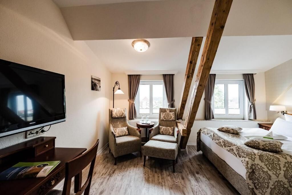 A hotel room with a TV, two armchairs, a bed, a small table, and a window with curtains.