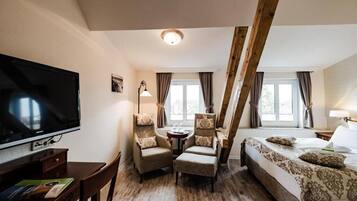 A hotel room with a TV, two armchairs, a bed, a small table, and a window with curtains.