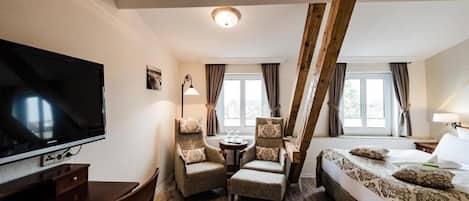 Hotellihuone, jossa on televisio, kaksi nojatuolia, sänky, pieni pöytä ja ikkuna, jossa on verhot.