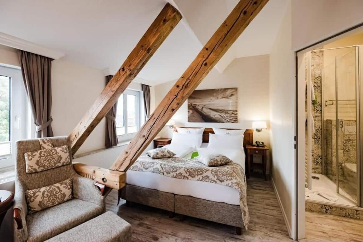 A bedroom with a sloped ceiling, a bed with patterned bedding, a chair with cushions, a bedside table, and a shower area.