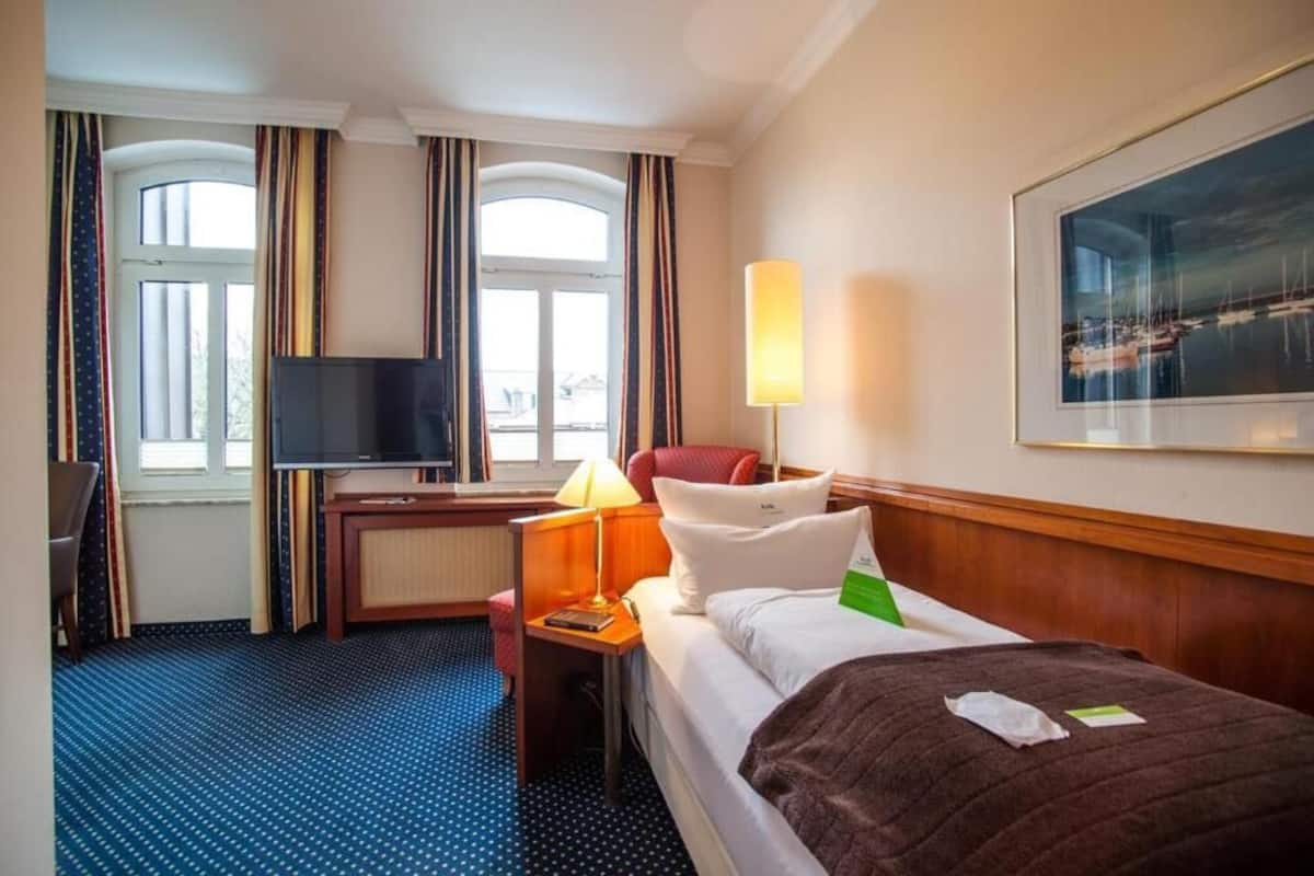 A hotel room with a bed, a desk, a TV, a lamp, and a painting on the wall.