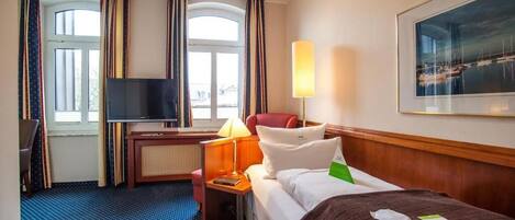 A hotel room with a bed, a desk, a TV, a lamp, and a painting on the wall.
