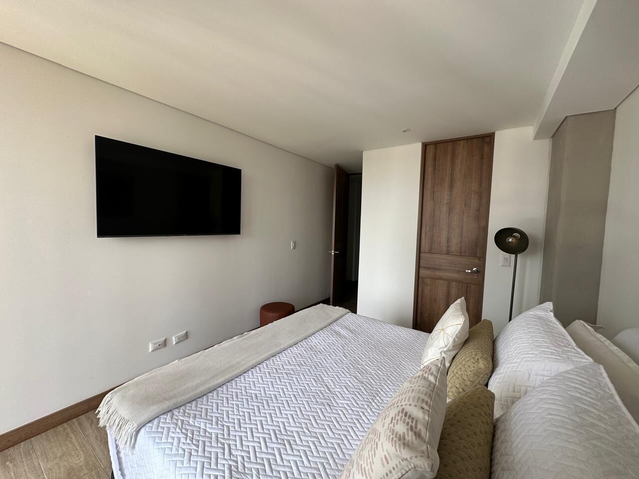 Une chambre à coucher comprenant un lit, un téléviseur à écran plat fixé au mur, une armoire en bois et une lampe sur pied.
