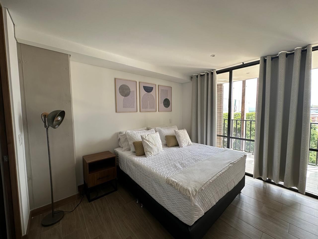 Un dormitorio con cama, mesita de noche, lámpara de pie y ventana con cortinas.