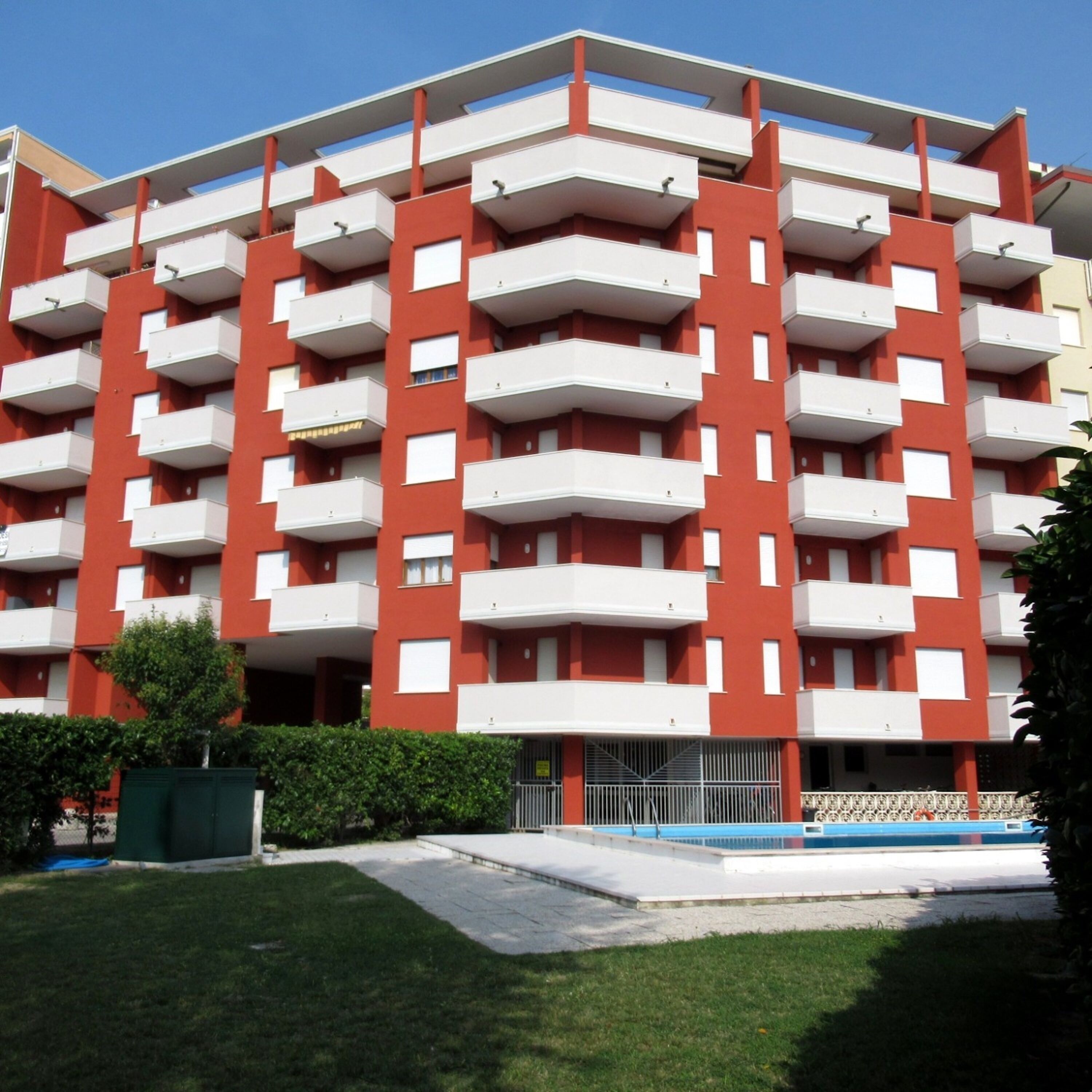 Ένα πολυώροφο κτίριο κατοικιών με κόκκινη και λευκή πρόσοψη, μπαλκόνια και μια πισίνα στο προσκήνιο.