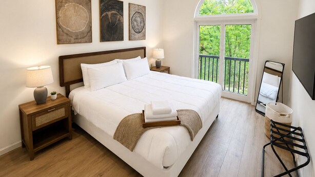 A bedroom with a large bed, a nightstand with a lamp, a bedside table with a lamp, a window with a view of greenery, and a wall-mounted TV. - The Accessible Atom | Historic Main (Saint Charles)