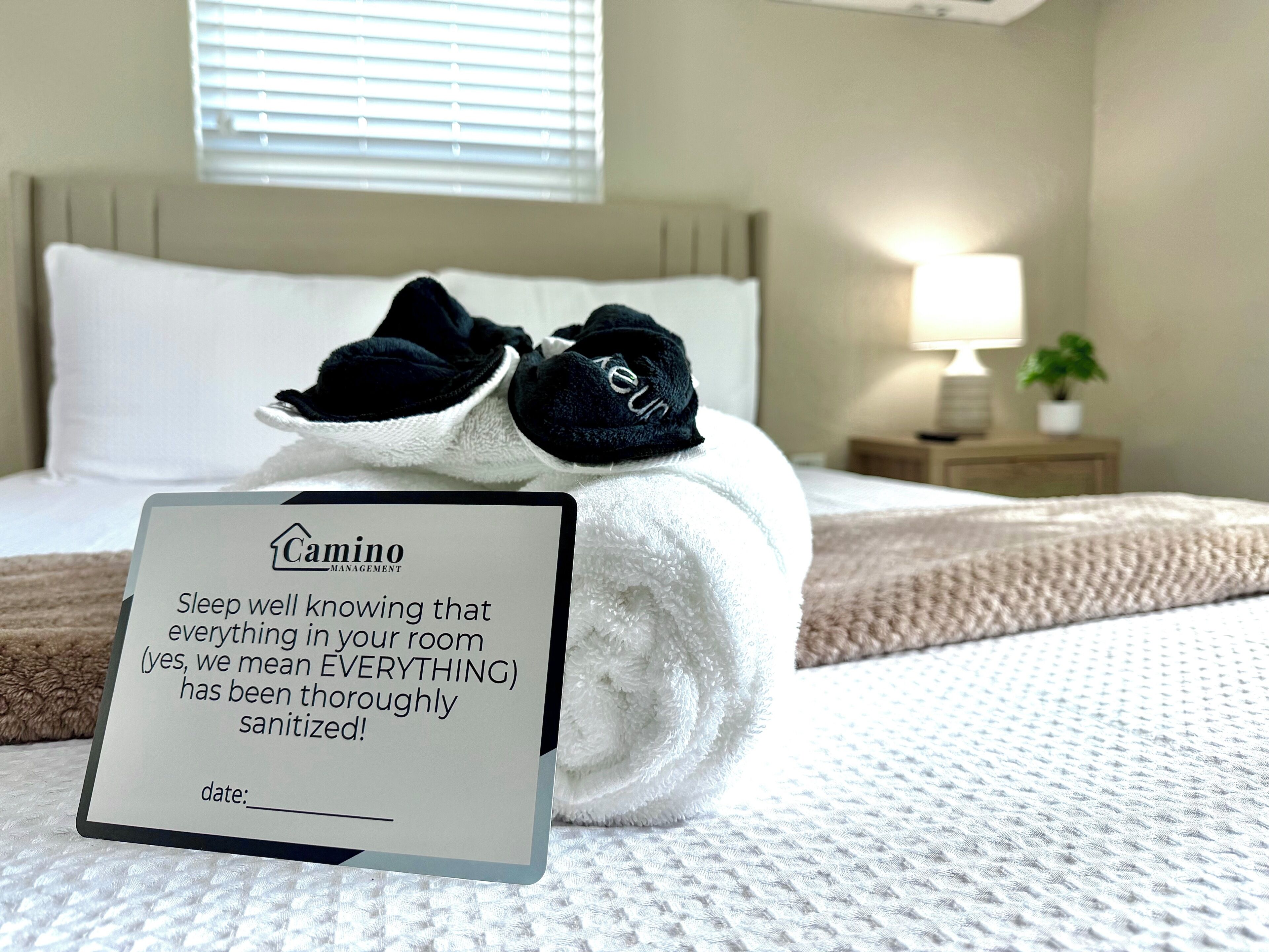 Un lit bien fait, avec un panneau indiquant que la chambre a été soigneusement désinfectée, un oreiller, une paire de chaussons, une lampe de chevet et une fenêtre avec des stores.
